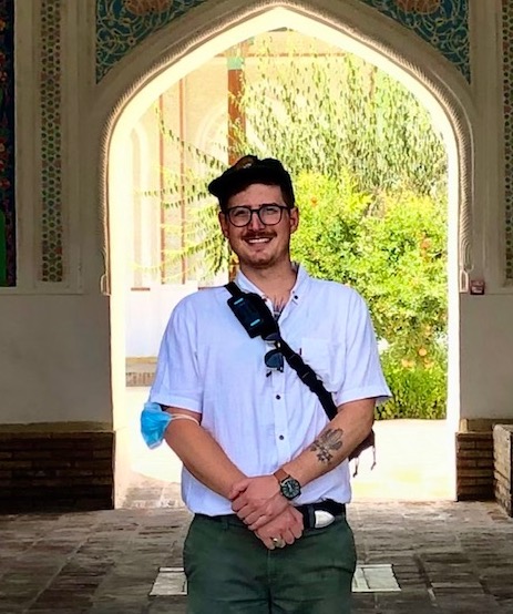 A close up shot of Matt Brown smiling in a large, ornate central asian doorway. He is a wearing a short-sleeve button-up, a baseball cap, and glasses.