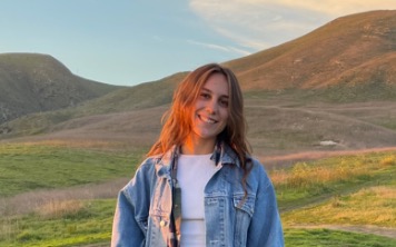 Photo of Nicole Wallius (brown hair, white shirt, jean jacket) standing in front of the Santa Barbara mountains. 