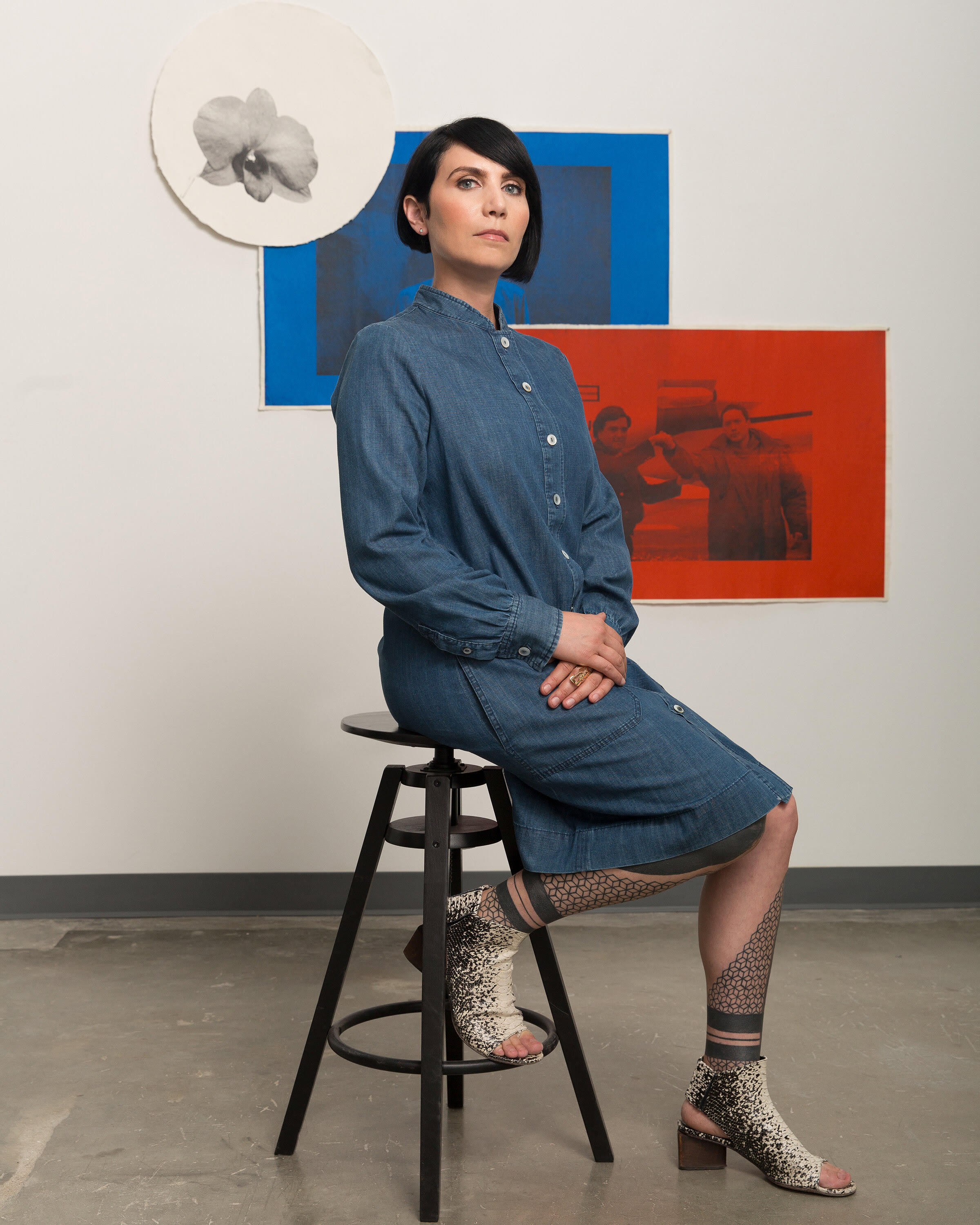 Regina Mamou, dressed in a blue dress with high heels, sits on a stool. In the background are two pieces of art. 